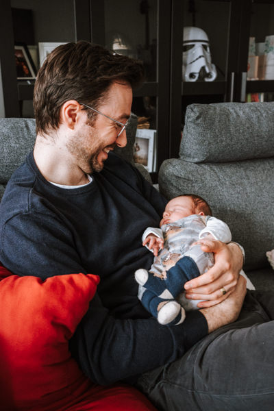 Papa sitzt auf grauer Couch und hält Baby im Arm während er stolz lacht