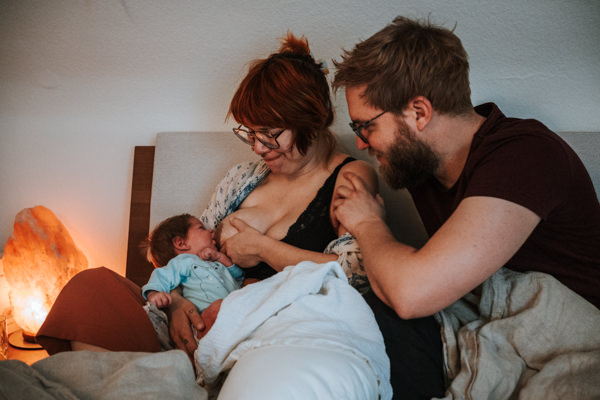 Familie im Wochenbett. Die Mama legt das Baby zum Stillen an. Dabei hat sie ein schmerzverzerrtes Gesicht. Der Papa sitzt nah bei ihr und hält ihre Schulter.