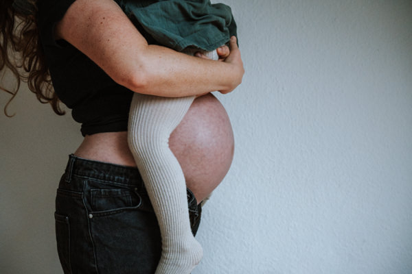 2. Nahaufnahme von der Seite. Schwangere Mama mit nacktem Bauch hat Kleinkind auf dem Arm. Man sieht von der Mama den Arm, den Bauch und vom Kleinkind die Beine in cremefarbener Strickstrumpfhose.