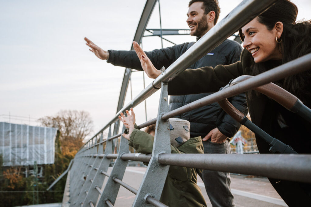 Familie steht auf einer Brücke. Mama und Papa sind zu erkennen, die Kindergesichter vom Geländer verdeckt. Sie schauen nach unten und winken den vorbeifahrenden Zügen. Die Züge sind nicht zu sehen.