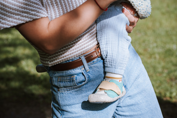 Detailaufnahme. Mama hat Kleinkind auf dem Arm. Man sieht den Arm von der Mama und Bein mit Fuß vom Kind. Im Hintergrund Wiese.