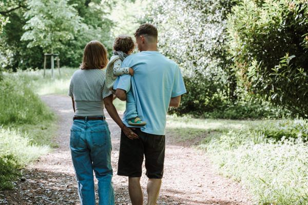 Im Park. Familie von hinten. Papa hat Kind auf dem Arm. Mama auf der anderen Seite vom Kind mit Hand am Gesäß vom Papa.