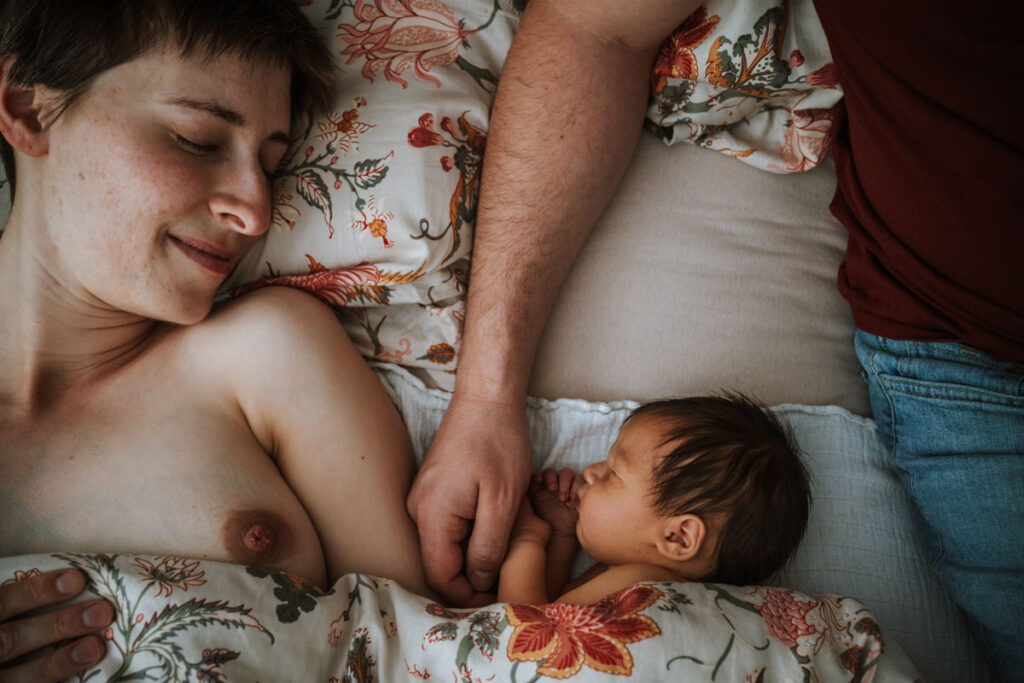 Familie liegt im Bett ein paar Tage nach der Geburt. Am unteren Bildrand sieht man die Bettdecke. Am linken Bildrand liegt die Mama. Man sieht ihre linke Brust und ihren Kopf, der zu den anderen gedreht ist. Sie lächelt mit geschlossenen Augen. Daneben liegt der Arm des Papas. Neben der Hand guckt der Kopf des Babys aus der Decke raus. Am rechten Bildrand sieht man noch den Körper des Papas zur Hälfte.