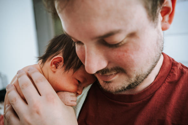 Nahaufnahme. Stolz lächelnder Papa hält Neugeborenes auf dem Arm an seiner Schulter. Beide gucken nach unten. Von dem Baby sieht man nur den Kopf. Der Rest des Körpers ist von der Hand des Vaters bedeckt.