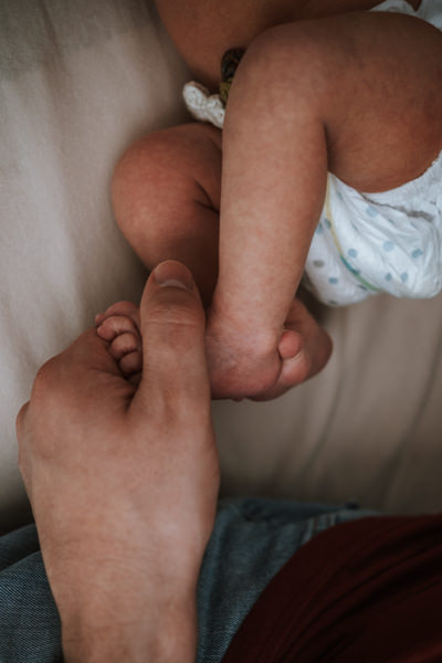 Detailaufnahme. Hochkantbild. Aus dem unteren Rand kommt die Hand des Papas, die die Füße des Neugeborenen hält, das bis zur Hüfte zu sehen ist.