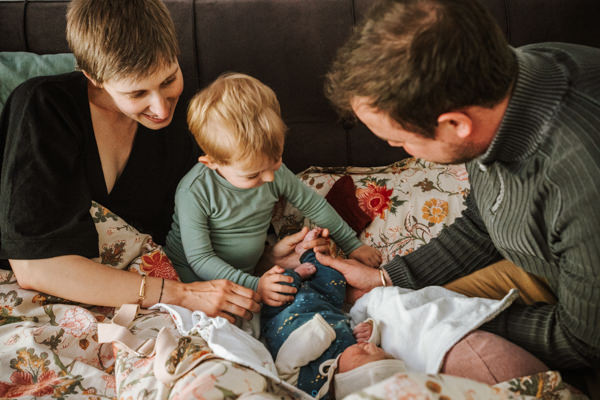 Eltern, Kleinkind und Neugeborenes im Bett. Halbe Vogelperspektive. Erstes Kennenlernen nach der Geburt. Kleinkind entdeckt Füße des Babys.