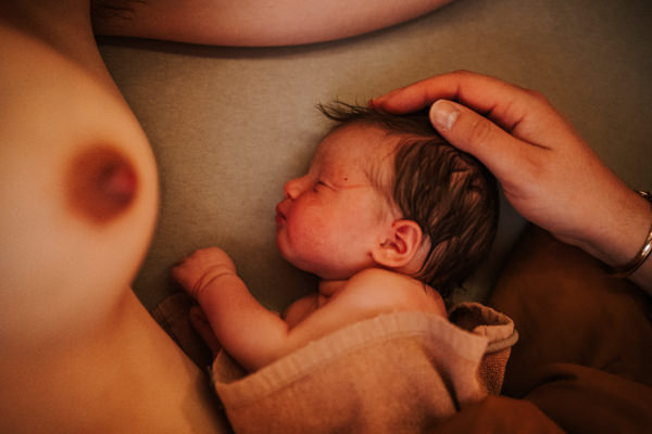 Frisch geborenes Baby liegt zwischen neben der nackten Brust der Mama mit der Hand des Papas auf dem Kopf