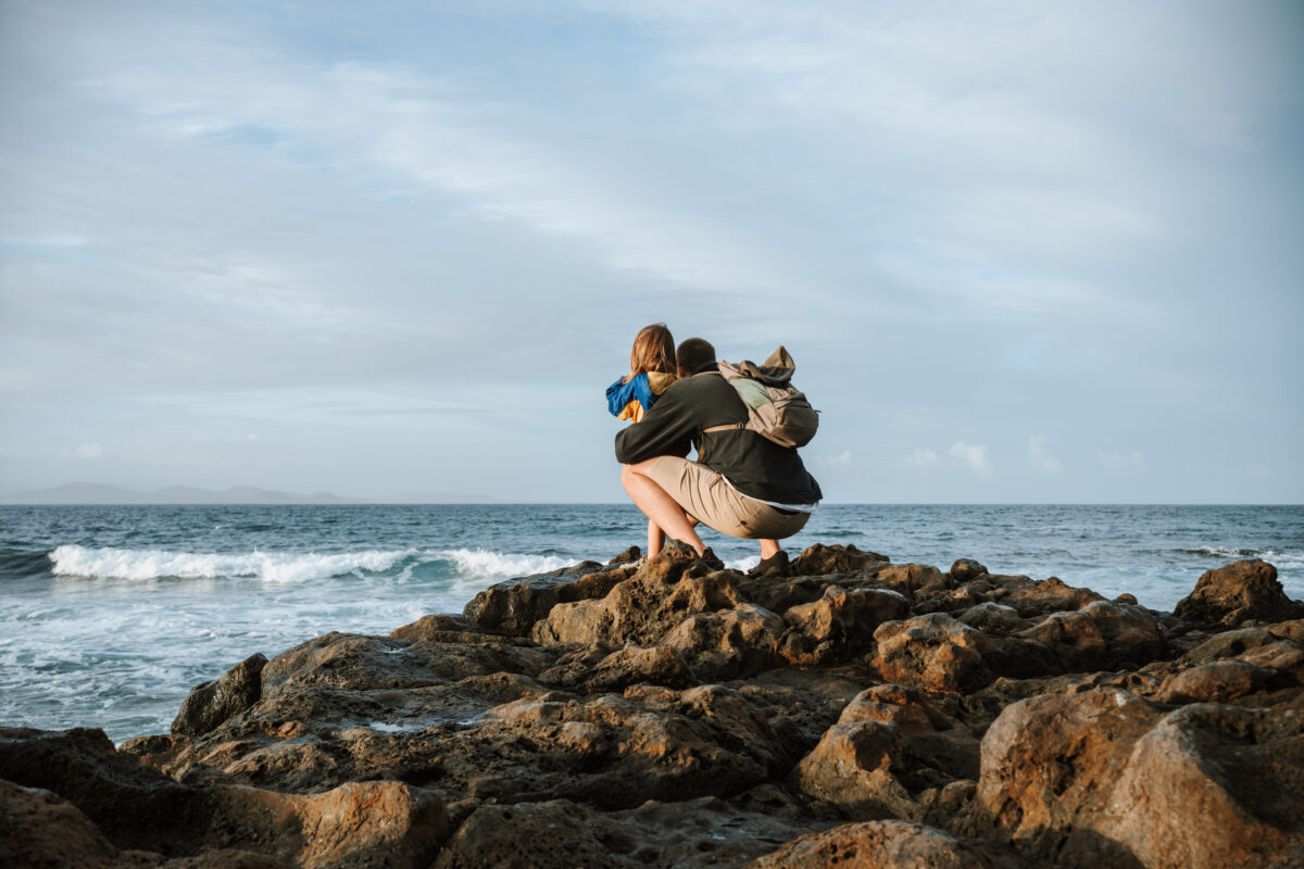Papa und Kind zum Sonnenaufgang auf Felsen am Meer mit dem Rücken zur Kamera. Der Papa hat einen Rucksack auf und sitzt in der Hocke. Das Kind steht vor ihm. Gemeinsam schauen sie in die Ferne.